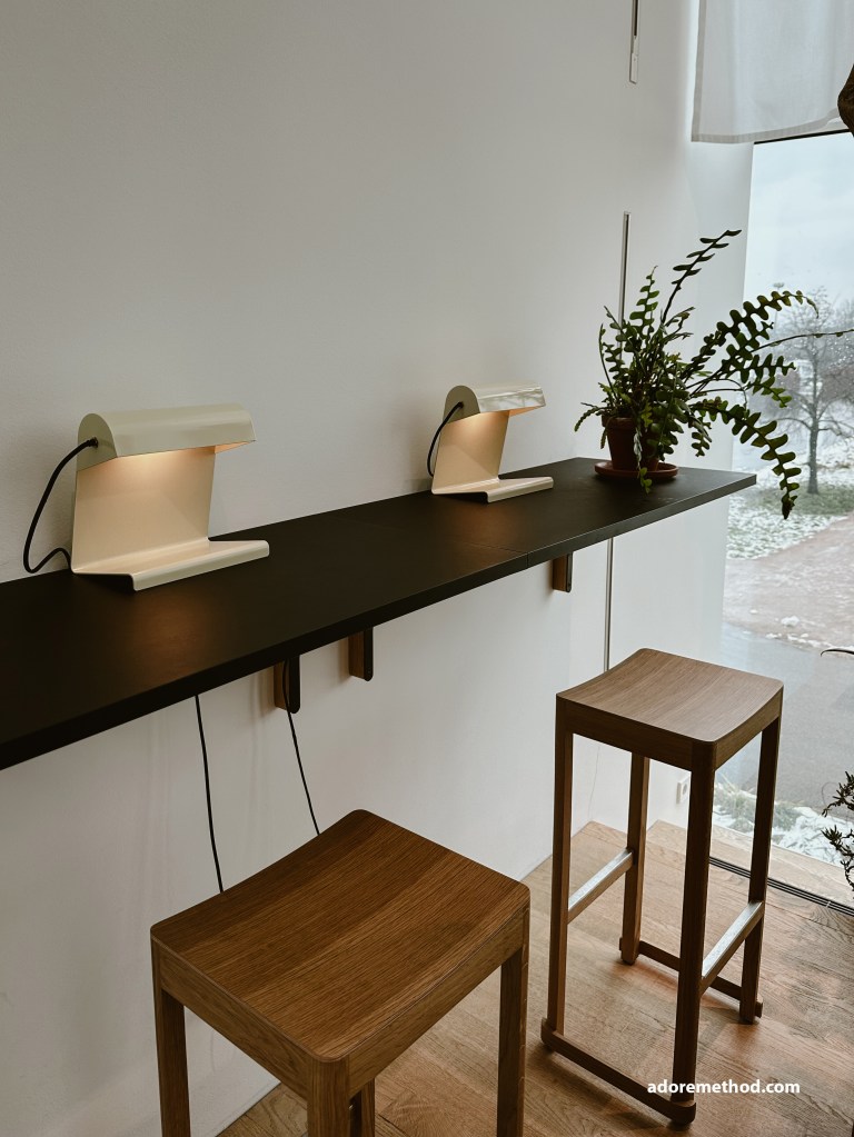 Un coin bar minimaliste avec un plan de travail noir suspendu, des tabourets en bois et deux lampes de bureau design, offrant un espace fonctionnel et lumineux avec vue sur l’extérieur.
