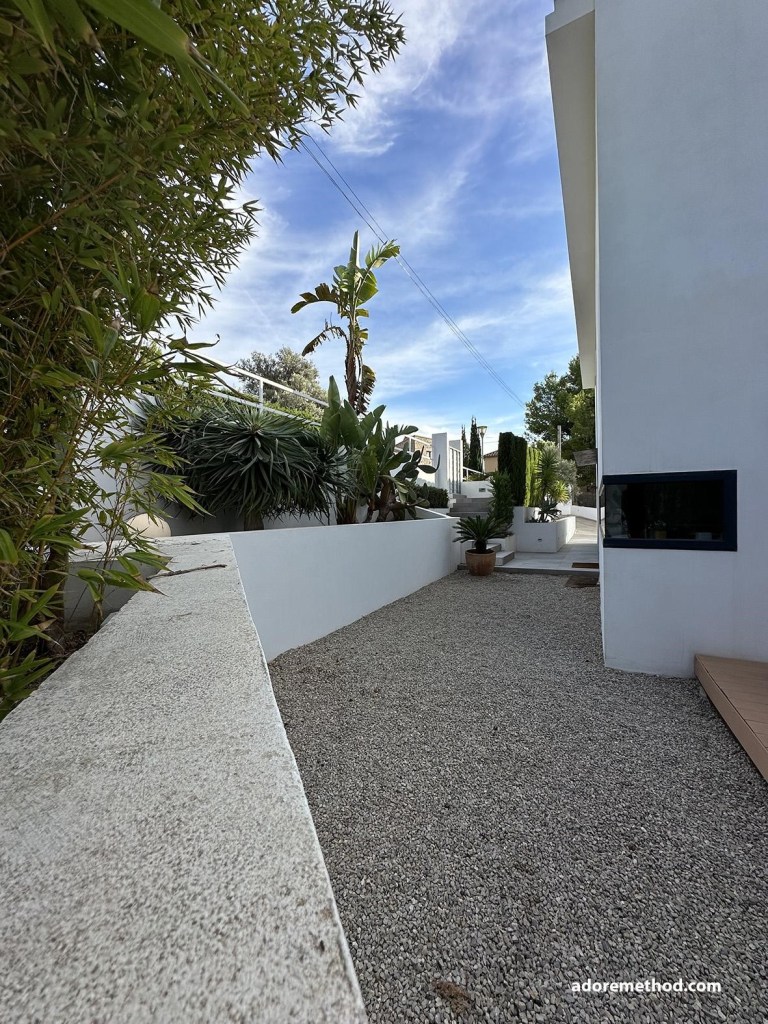 Vue d'une allée extérieure gravillonnée bordée de bambous et de plantes vertes devant une maison moderne.
